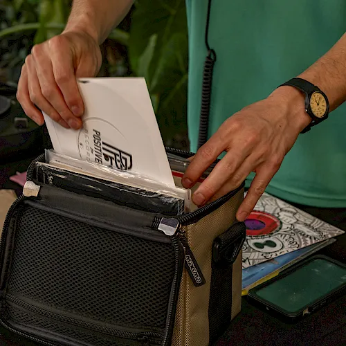 A person in a green shirt places printed mail/envelopes into a zippered organizer pouch outdoors, with a watch on their wrist.