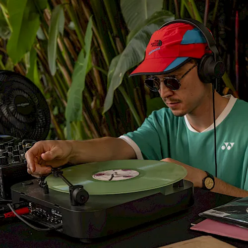 A man in headphones and a cap DJs at a turntable with vinyl records, surrounded by tropical plants and colorful sleeves.