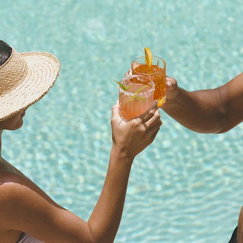 Two people by a pool clink glasses with colorful drinks, enjoying a sunny day.