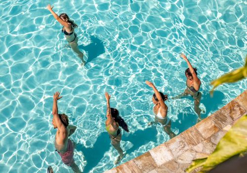 F&uuml;nf Personen trainieren in einem Schwimmbecken und f&uuml;hren synchrone Bewegungen am Beckenrand aus.