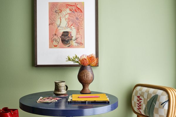 A cozy corner with a round table, decorative vase, mug, chair, and framed art on a light green wall.