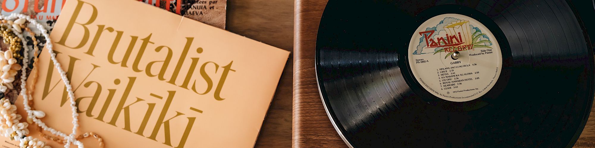 A vinyl record on a wooden turntable, with album covers, a beaded necklace, and "Brutalist Waikiki" visible.
