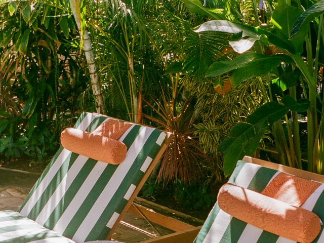 Two striped lounge chairs with orange pillows sit under a purple umbrella, surrounded by lush greenery and a small round table.