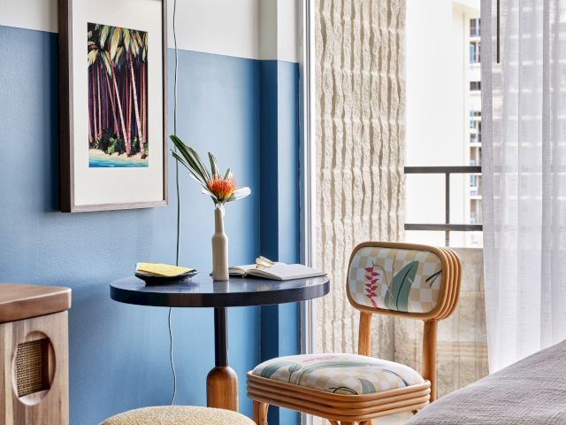 A cozy room with a blue accent wall, framed tropical print, colorful hanging lamp, small table, floral arrangement, and a woven chair.