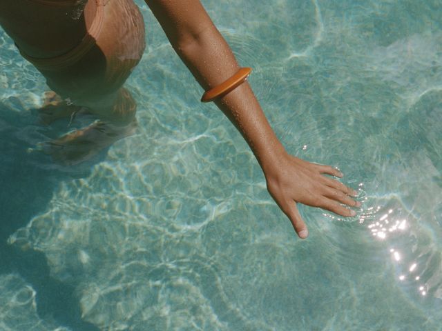 A person in a swimsuit stands in clear, sunlit water with an orange bracelet on their wrist, their hand touching the water surface.
