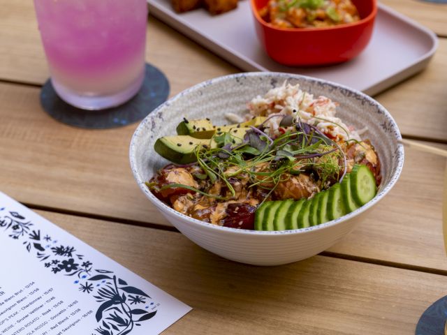A bowl with a salad, pink drink, skewered food, and a red bowl with dip are on a wooden table, with a menu on the side.