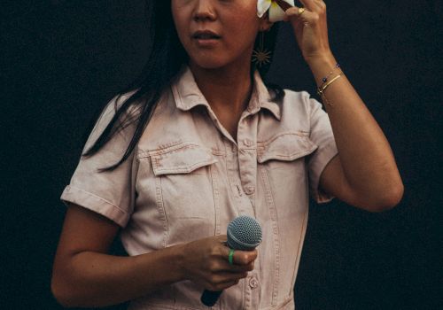 A person in a light outfit holds a microphone and adjusts a flower in their hair against a dark background.