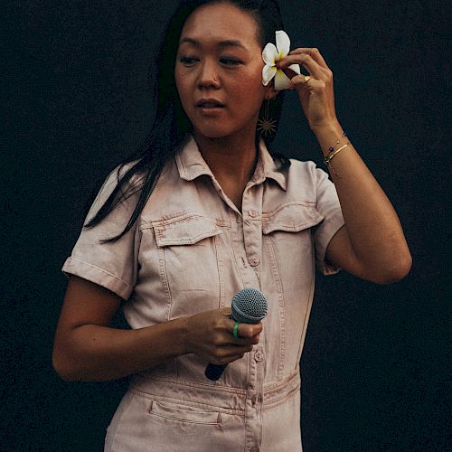 A person in a light outfit holds a microphone and adjusts a flower in their hair against a dark background.