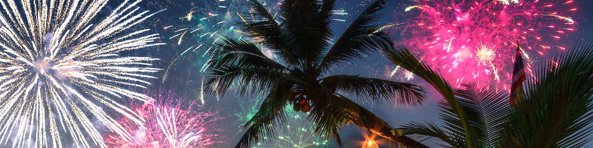 A tropical beach scene at sunset features palm trees, tiki torches, and vibrant fireworks lighting up the night sky.