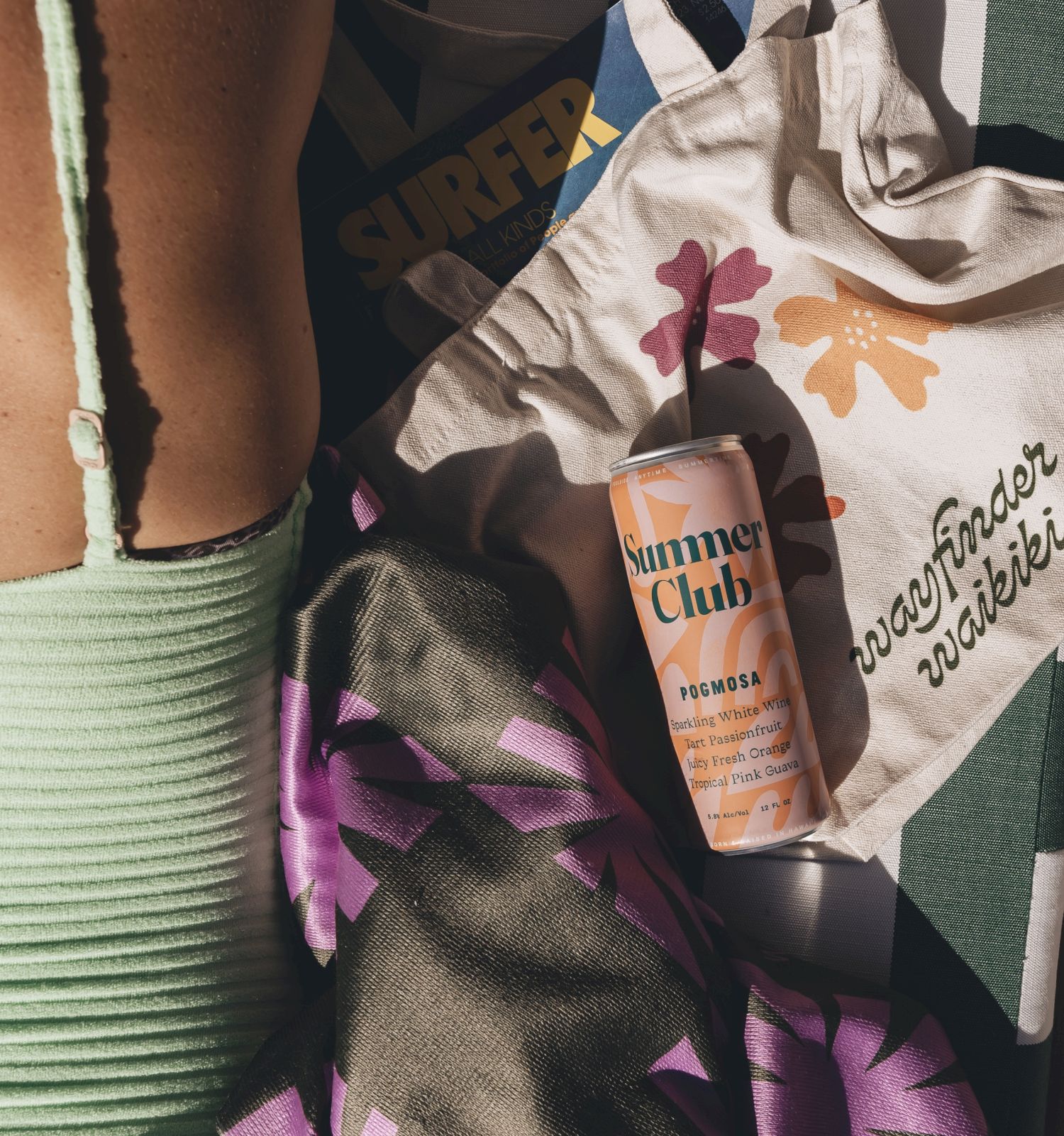 A person in a green swimsuit, a colorful towel, a can, a tote bag with floral design, and a magazine titled "SURFER" are visible.
