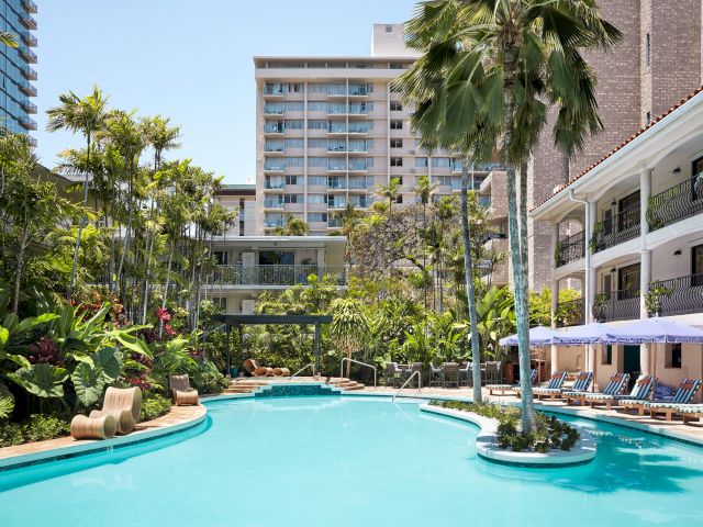 The image depicts a tropical resort-style pool area surrounded by lush greenery, modern buildings, and striped lounge chairs under umbrellas.
