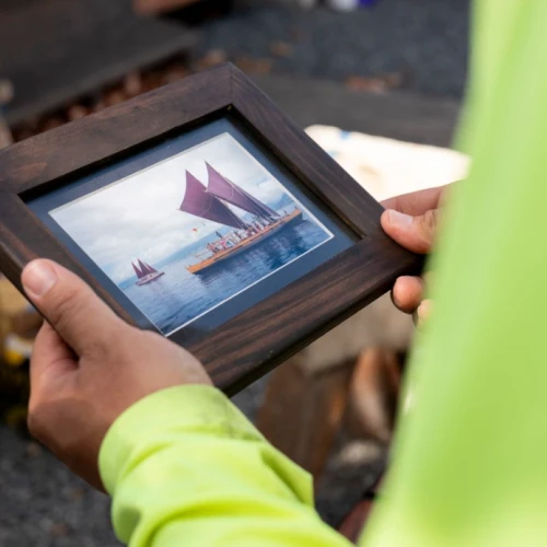 A person in a yellow sleeve holds a framed photo of a sailboat with red sails on the water.