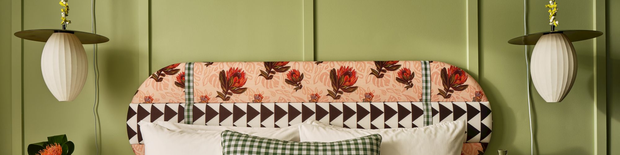 A cozy bedroom with a floral and geometric headboard, green checkered pillow, hanging lamps, and green paneled wall, creating a warm ambiance.