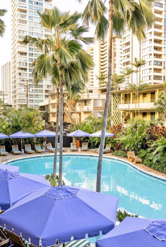 A pool area with blue umbrellas and palm trees is surrounded by tall buildings and lush greenery.