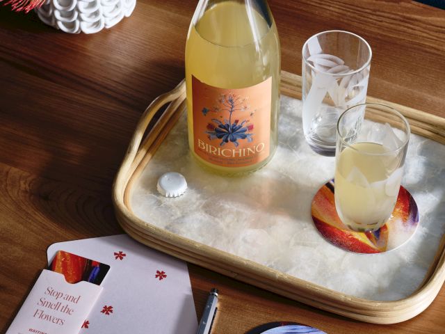 A bottle and glass on a tray with decorated coasters, a pen, and an open booklet on a wooden table with a floral arrangement.