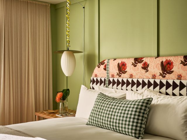 A cozy bedroom with green walls, a floral headboard, and a checkered pillow, featuring soft lighting and beige curtains.