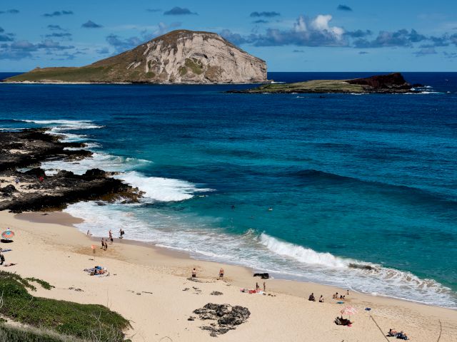 A beautiful beach scene with turquoise water, sandy shore, and people relaxing. There's an island visible in the distance under a bright blue sky.