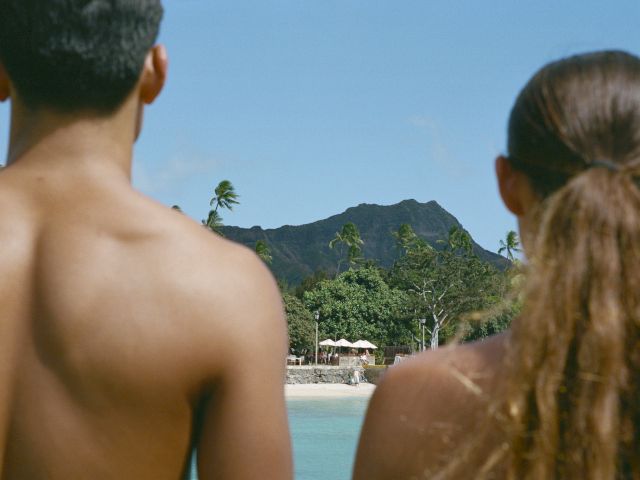 A shirtless man and woman facing a scenic view of trees, mountains, and clear sky on a beach.