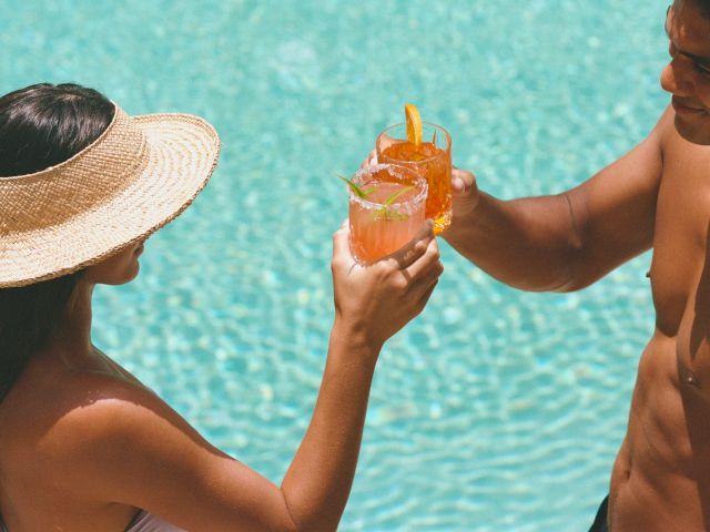 Two people by a pool are clinking glasses filled with colorful drinks. One is wearing a sun hat; both appear relaxed and enjoying the moment.