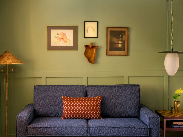 A cozy living room with a blue sofa, green walls, art, wooden tables, and a warm light fixture.