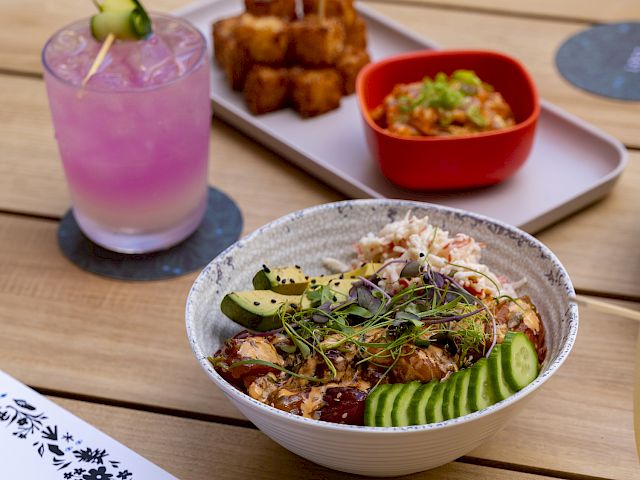 A table with a pink drink, a bowl of salad with cucumbers, a tray of skewered snacks, and a red bowl with a side dish, plus a paper menu.