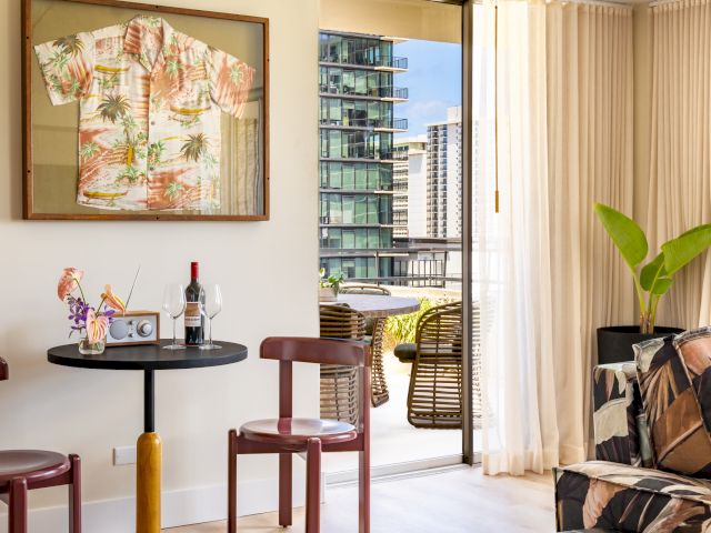 A room with a framed shirt, table with wine, chairs, sofa, plant, and city view through open balcony doors.