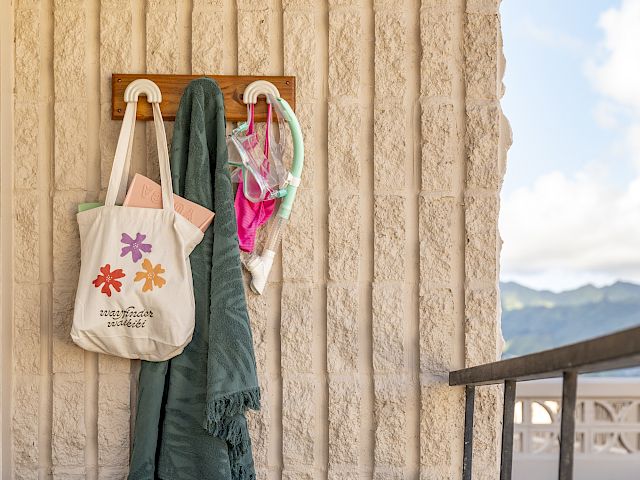 A cozy balcony with a rattan chair, a table with a mug, a tote bag on a hook, and a view of mountains in the background.