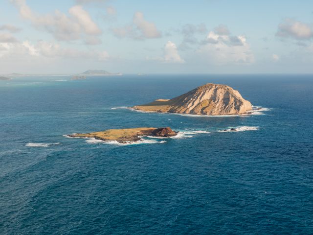 An island in the ocean with a rugged hill, surrounded by blue water and scattered clouds in the sky.
