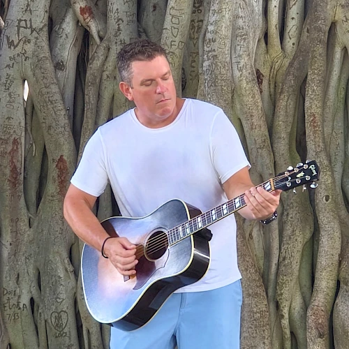 A person is playing a guitar in front of a large tree with exposed roots, which have various engravings on them.