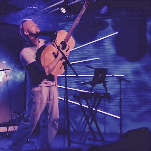 A musician performs passionately with an acoustic guitar on stage, surrounded by vibrant blue lighting and a keyboard stand.