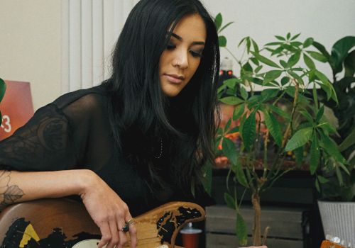 A person with long dark hair is sitting and playing an electric guitar in a room with plants in the background.