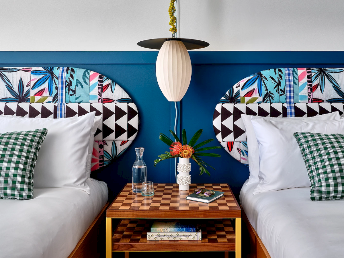 A colorful bedroom with two beds, checkered pillows, a geometric headboard, and a central nightstand with a lamp, books, and flowers.