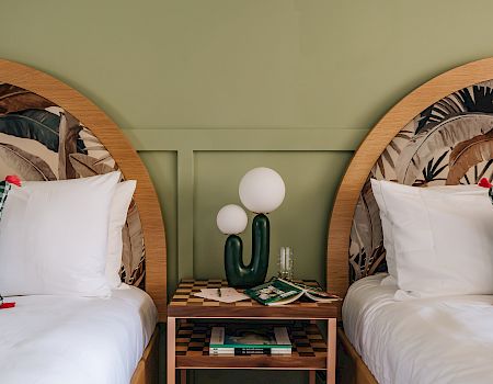 Two twin beds with patterned headboards, green accent wall, checked pillows. A cactus-shaped lamp and books are on a shared bedside table.