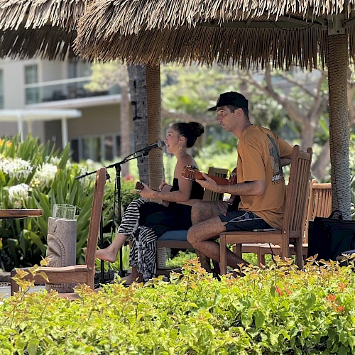 A man and woman are playing guitars and singing under a thatched roof gazebo surrounded by greenery.
