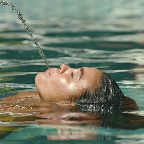 A person floating on their back in a pool and playfully spouting a stream of water upwards from their mouth, captured in clear detail.