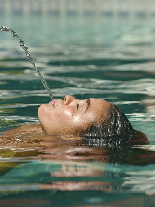 A person floating on their back in a pool and playfully spouting a stream of water upwards from their mouth, captured in clear detail.