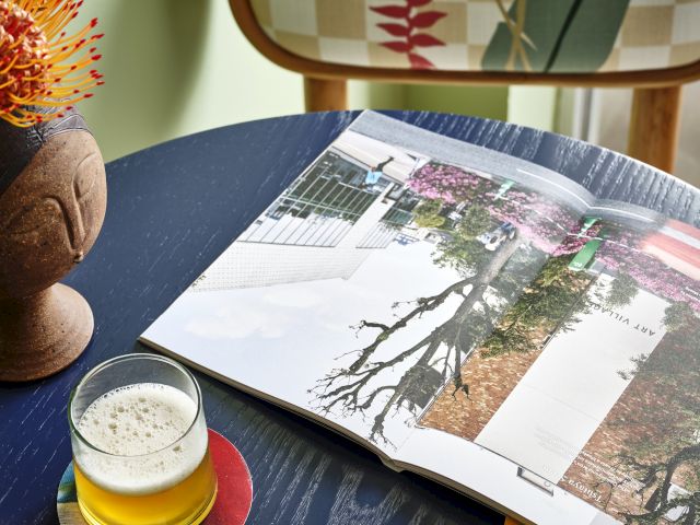 A cozy scene with an open book, a drink on a coaster, and pink floral postcards on a dark table, with a patterned chair in the background.