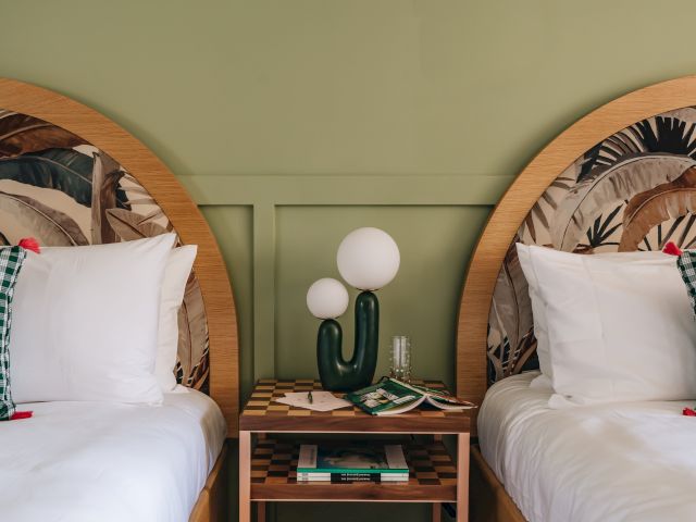 The image shows two beds with patterned headboards, a shared nightstand, and a cactus-shaped lamp, against a green wall.