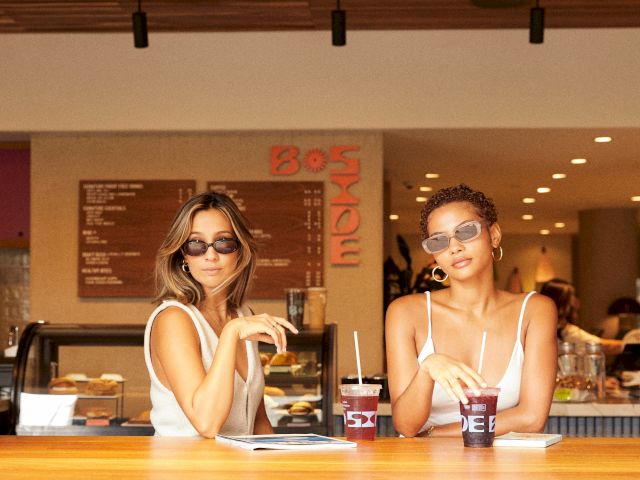 Two people wearing sunglasses sit at a counter with drinks in a cafe setting. The background includes a menu board and interior details.
