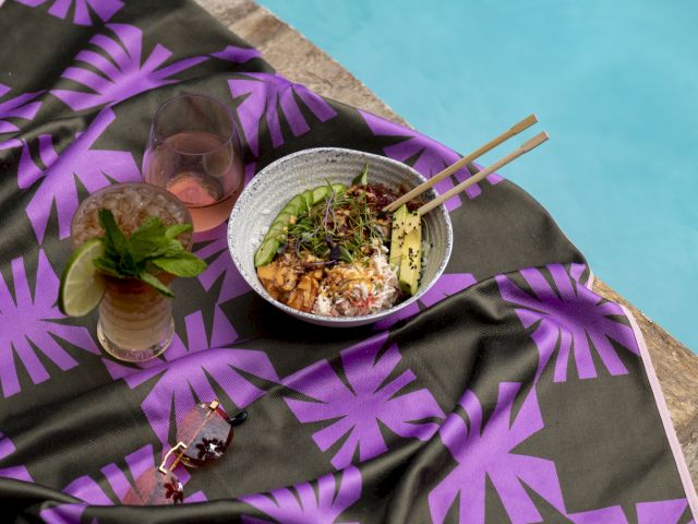 A bowl of food with chopsticks and a drink sit on a purple-patterned cloth by a pool. Sunglasses are also placed nearby.