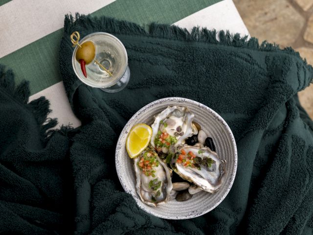 A bowl of oysters with lemon and garnish sits on a dark cloth next to a martini with an olive and a cocktail pick.