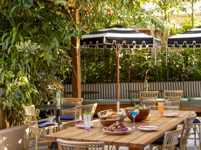 A cozy outdoor dining area with wooden tables, chairs, plants, and black umbrellas. Plates and glasses are set for a meal.