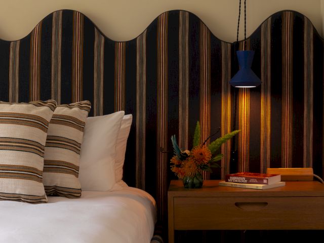 A cozy bedroom setup with a striped headboard, pillows, a bedside table, lamp, books, and flowers.