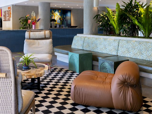 The image shows a stylish lounge area with modern furniture, plants, and a checkered floor design, creating a cozy atmosphere.