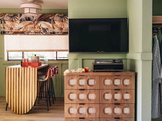 The image shows a room with green walls, a wall-mounted TV, a striped cabinet, a small kitchen counter with chairs, and patterned curtains.