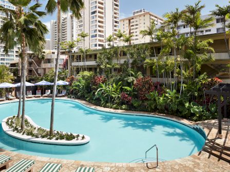 Two people in swimwear stand partially hidden by green foliage, creating a playful and summery scene.