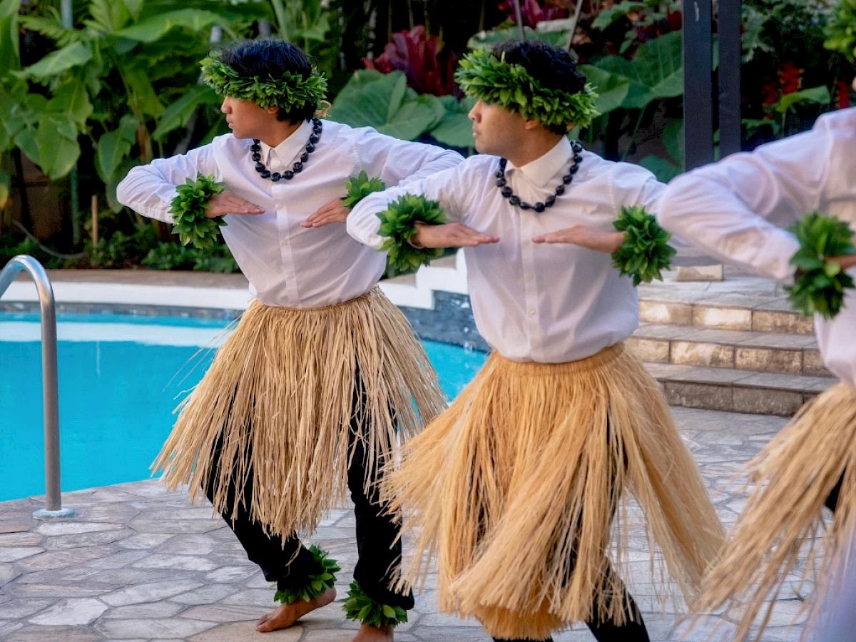 Dancers in traditional attire perform near a swimming pool, wearing grass skirts and greenery accessories, surrounded by lush plants.