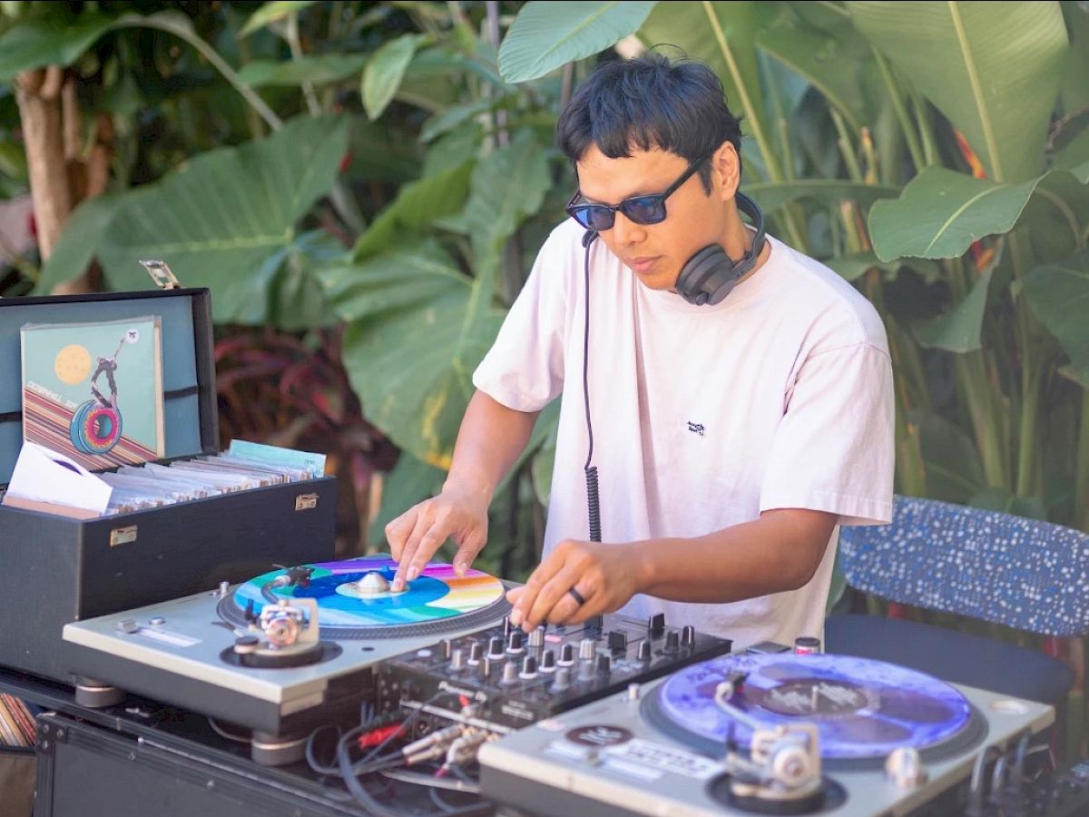 A DJ wearing sunglasses and a T-shirt is mixing music on a turntable setup outdoors, surrounded by lush green plants and trees ending the sentence.