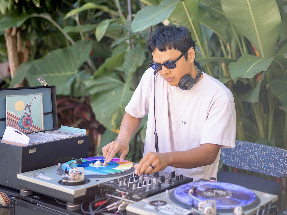 サングラスをかけたDJが、熱帯植物や機材に囲まれた屋外でターンテーブルを回し、ヘッドホンを装着して楽曲をミックスしている。