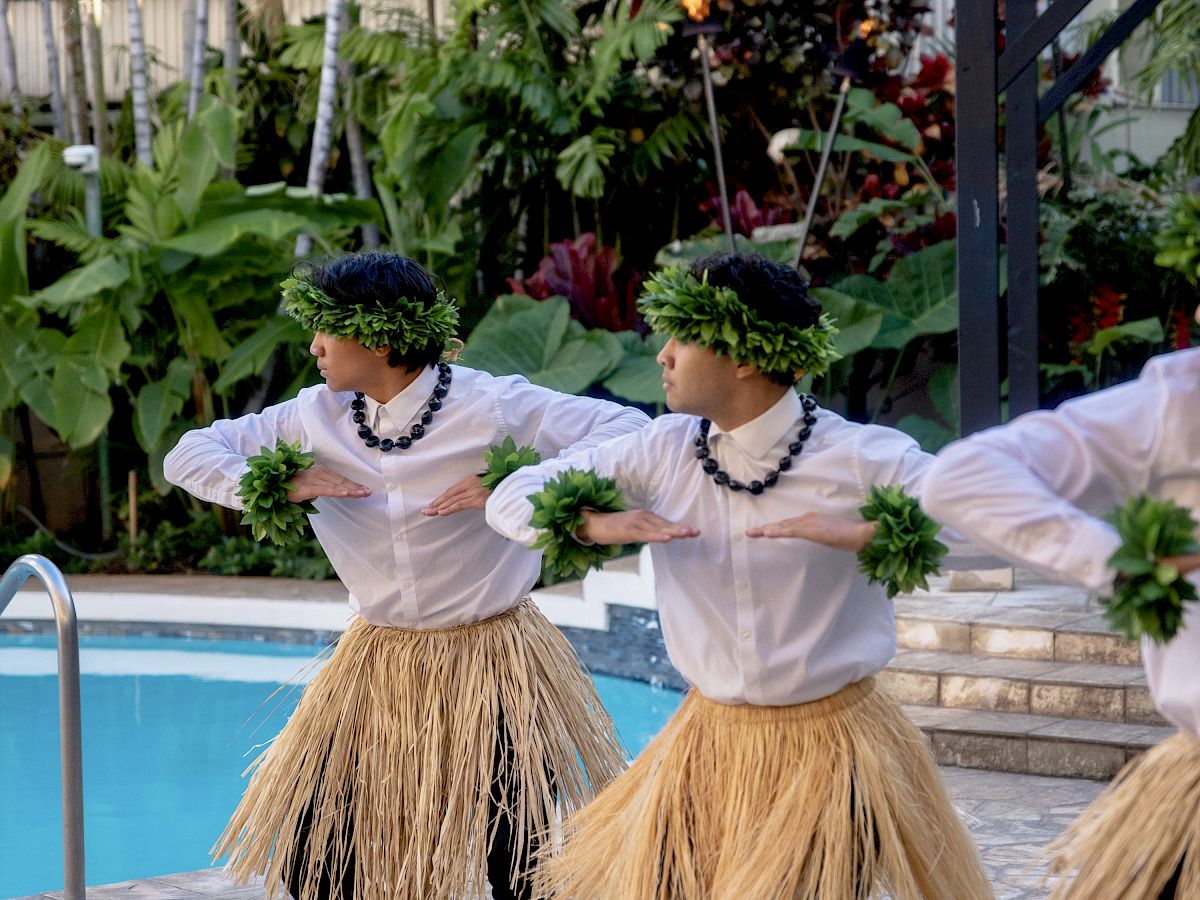 草のスカートとレイを身に着けた3人が、緑の花束を手に、熱帯の屋外のプールサイドでポリネシアのフラダンスを踊っている。